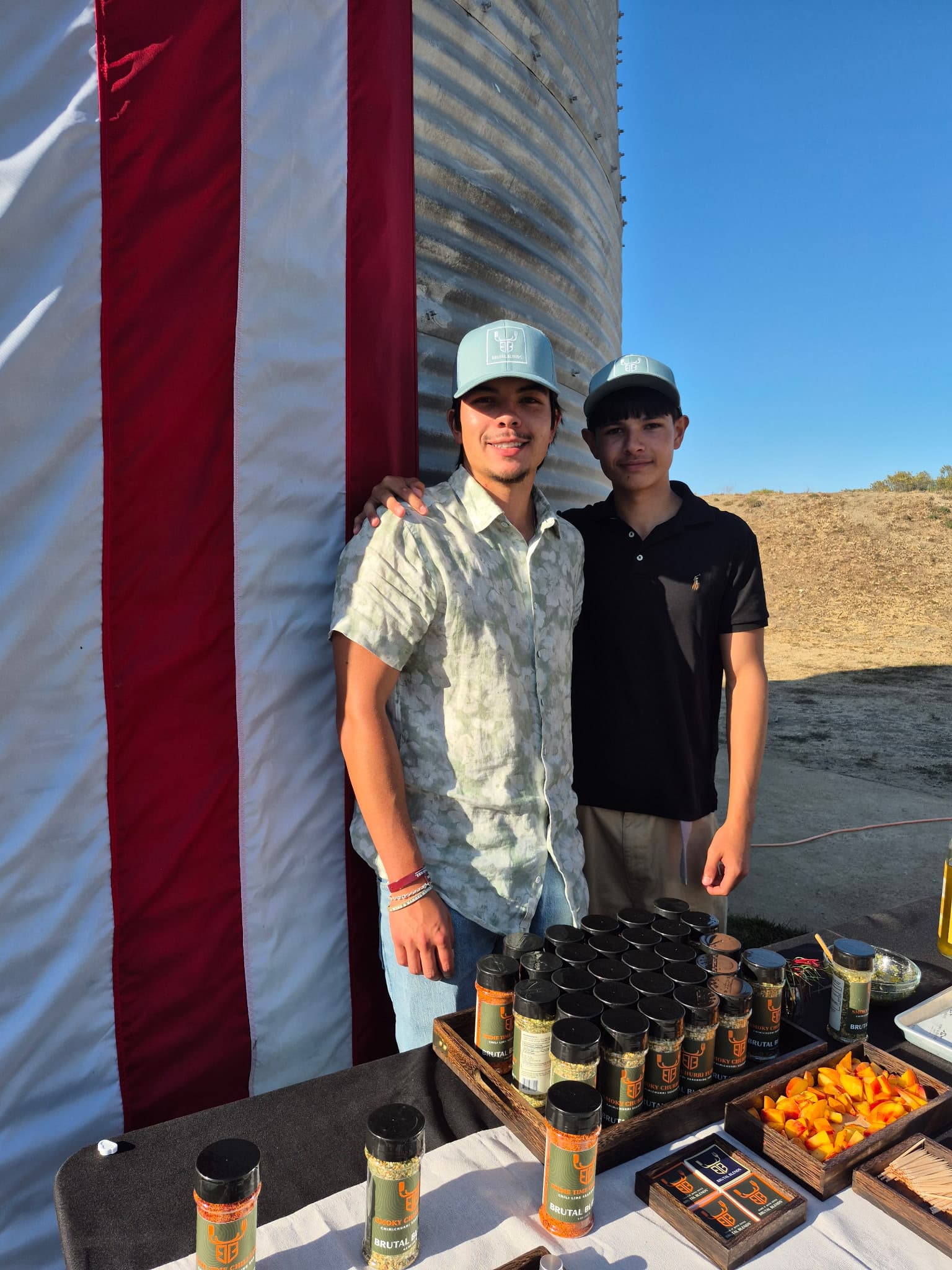Two men in blue hats stand behind a table of spices next to a silo.