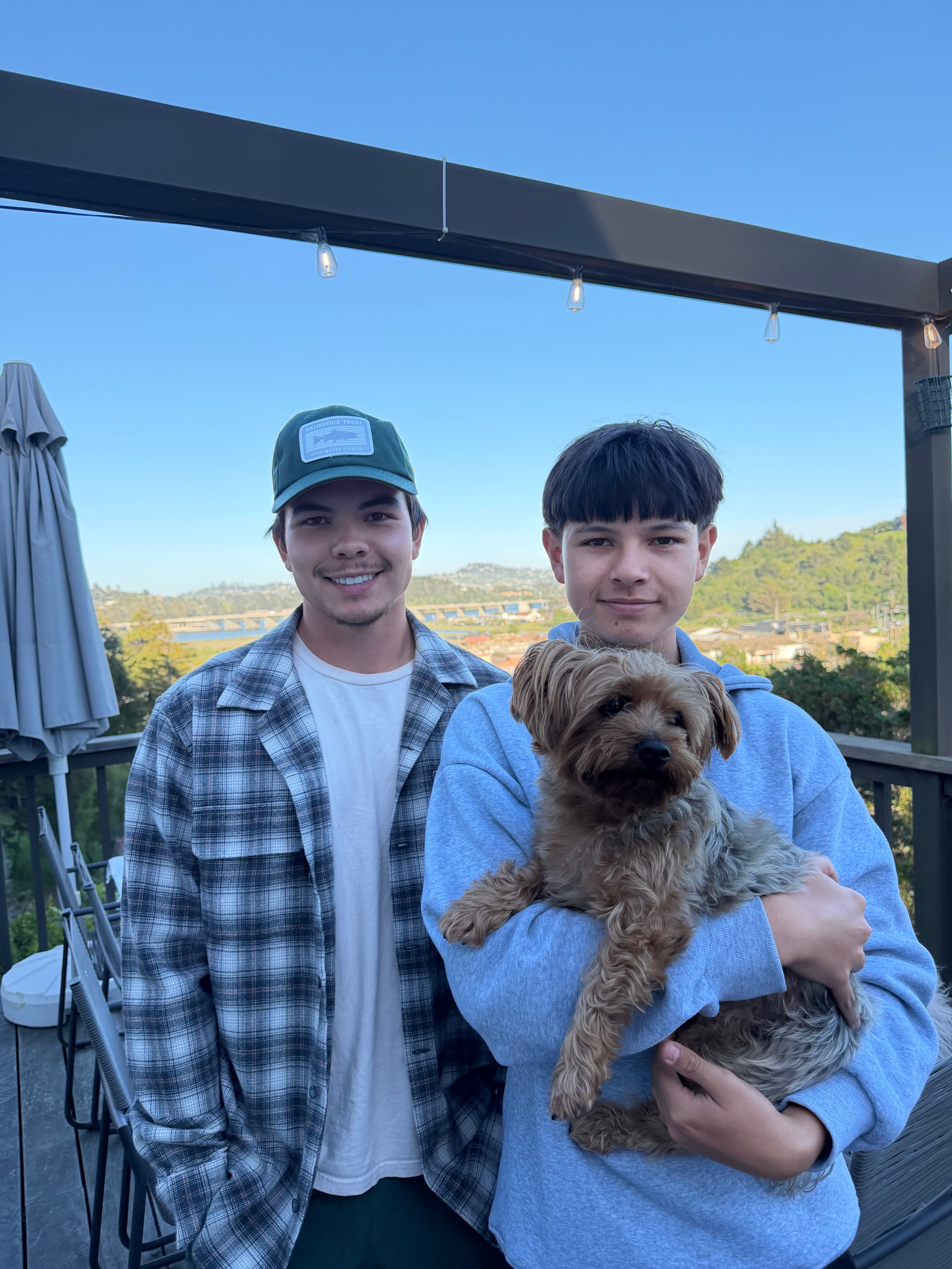 Two young men stand on a deck, one holding a small dog, with hills behind.